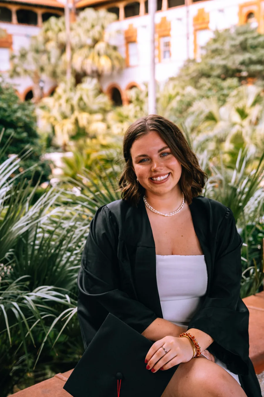 Sophie Bowman smiling in a graduation cap and gown, seated outdoors among palm trees
