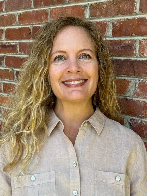 Staci Harrub smiling outdoors in front of a brick wall, wearing a tan button-up shirt