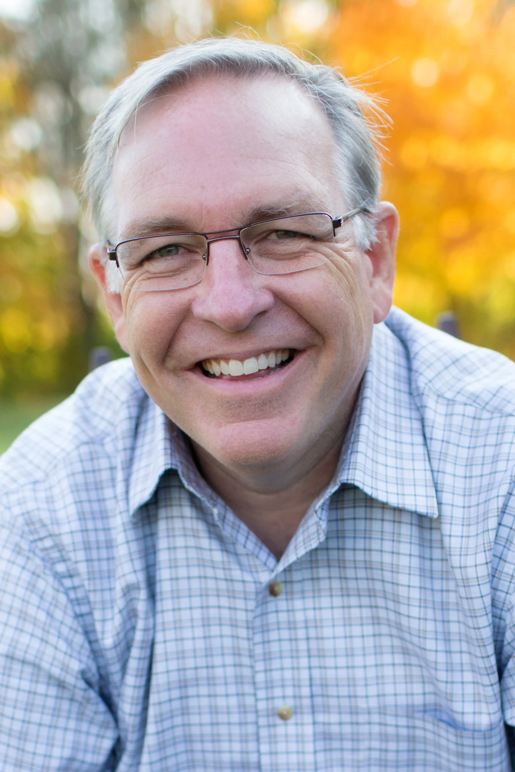 Tony Gore smiling outdoors in a plaid shirt, with autumn foliage in the background